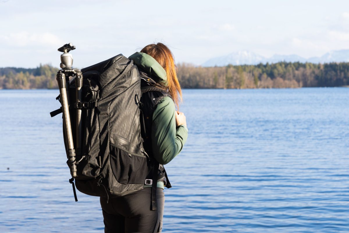 f-stop Ambassador Pia Steen wearing the f-stop Shinn 80 liter DuraDiamond® camera backpack with a tripod attached and standing near a lake with mountains in the background