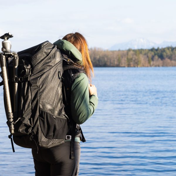 f-stop Ambassador Pia Steen wearing the f-stop Shinn 80 liter DuraDiamond® camera backpack with a tripod attached and standing near a lake with mountains in the background