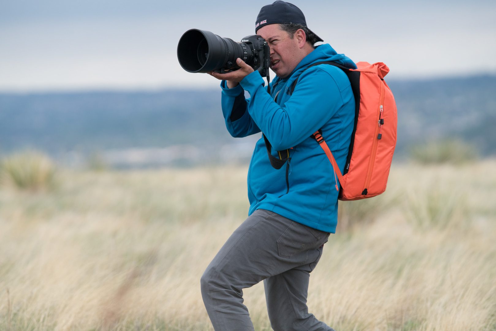 f-stop Ambassador David Clifford wearing the f-stop Dalston Urban Series 21 liter camera backpack in the Nasturtium color option and holding a Nikon D5 DSLR and a Nikon 200mm lens