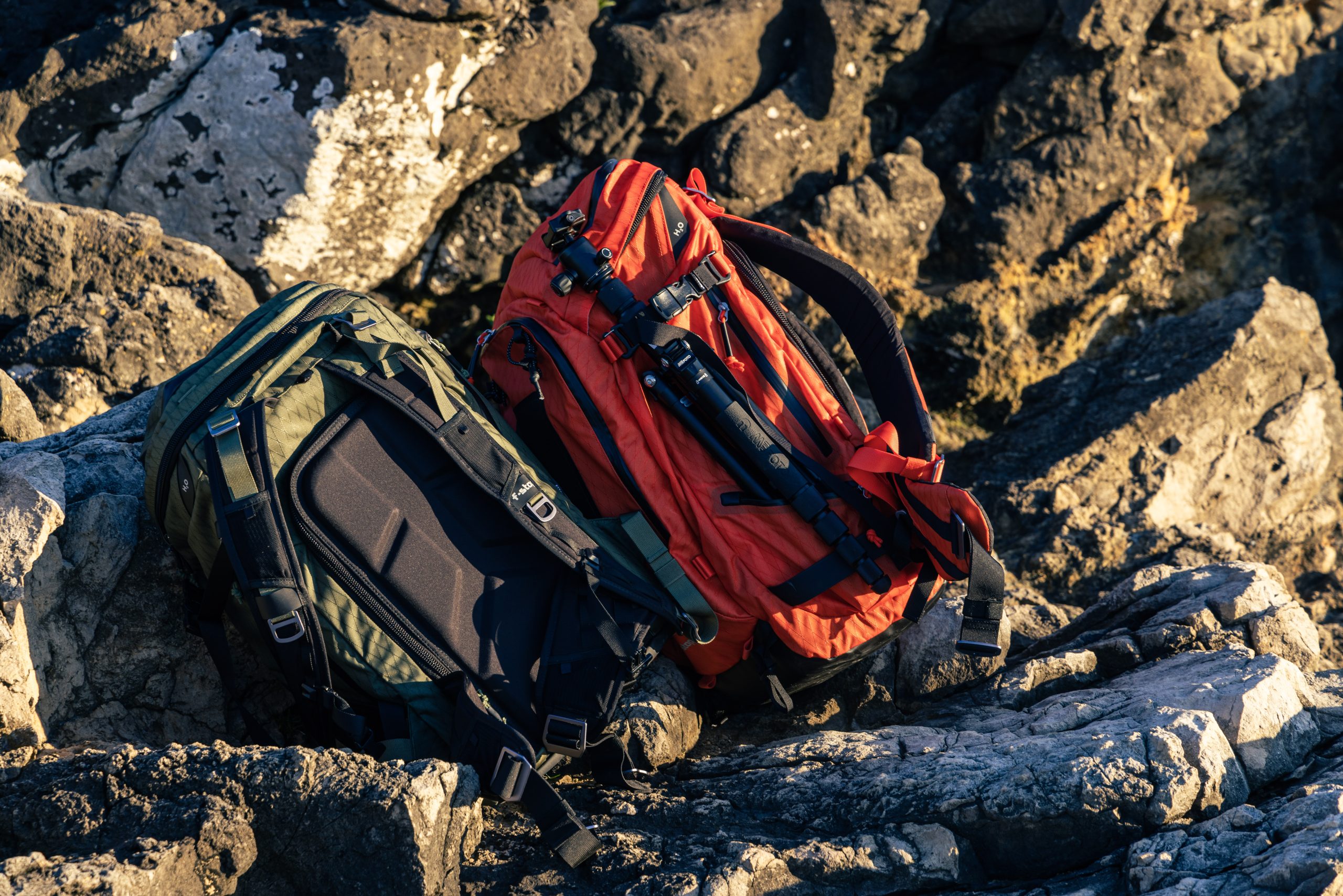 f-stop AJNA 37L camera backpack in Cypress Green color and the f-stop TILOPA 50L camera backpack in Magma Red color pictured on rocks